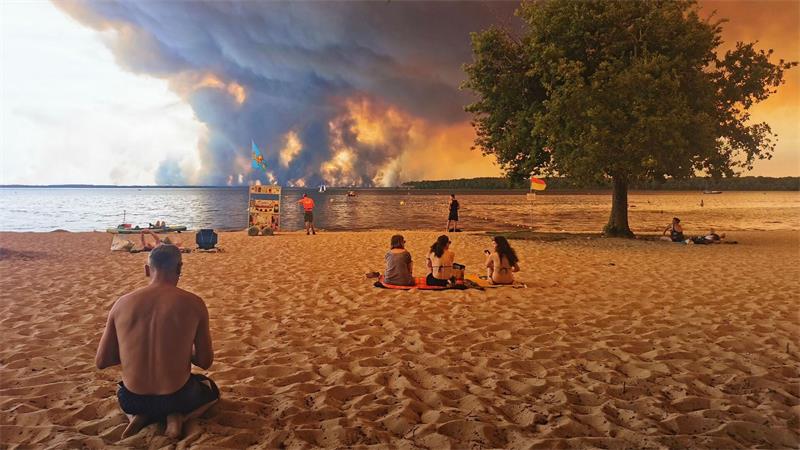 Strandbesucher sitzen am Strand von Moutchic in Lacanau im Südwesten Frankreichs, während Rauchwolken von einem Waldbrand in den Himmel aufsteigen.