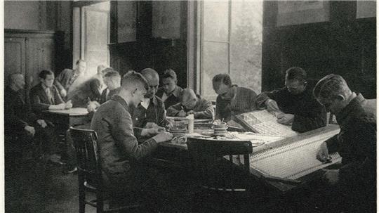 Mehrere Männer sitzen an Tischen in einem Raum und arbeiten konzentriert mit Papier und Werkzeugen