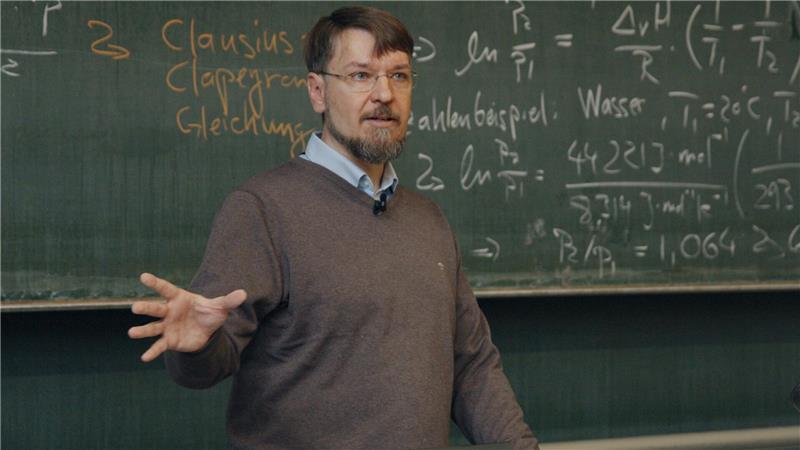 Ein Dozent an der Universität gestikuliert mit seiner rechten Hand. Er trägt eine Brille.