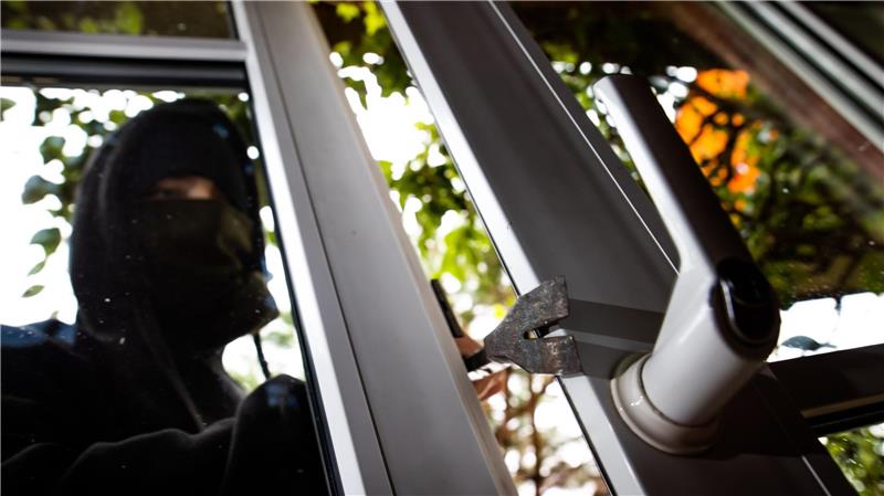 Eine in schwarz gekleidete Person dringt mit einer Brechstange durch ein Fenster in ein Gebäude ein.