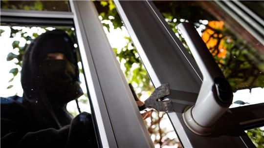 Eine in schwarz gekleidete Person dringt mit einer Brechstange durch ein Fenster in ein Gebäude ein.