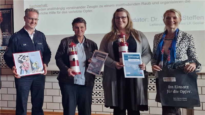Vier Personen stehen nebeneinander vor einer Wand mit Textprojektion und halten jeweils Urkunden, Feuerlöscher oder eine Tasche mit Aufschrift