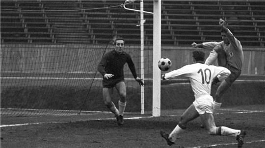 Schwarz-weiß-Foto einer Fußballszene mit Torwart, der sich auf einen Schuss eines Spielers mit Trikotnummer 10 vorbereitet, während ein Verteidiger versucht, den Ball abzuwehren