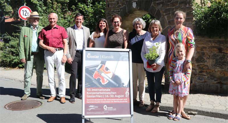Gruppe von neun Personen unterschiedlichen Alters steht im Freien neben einem Plakatständer mit Ankündigung für ein Konzertfestival