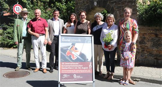 Gruppe von neun Personen unterschiedlichen Alters steht im Freien neben einem Plakatständer mit Ankündigung für ein Konzertfestival