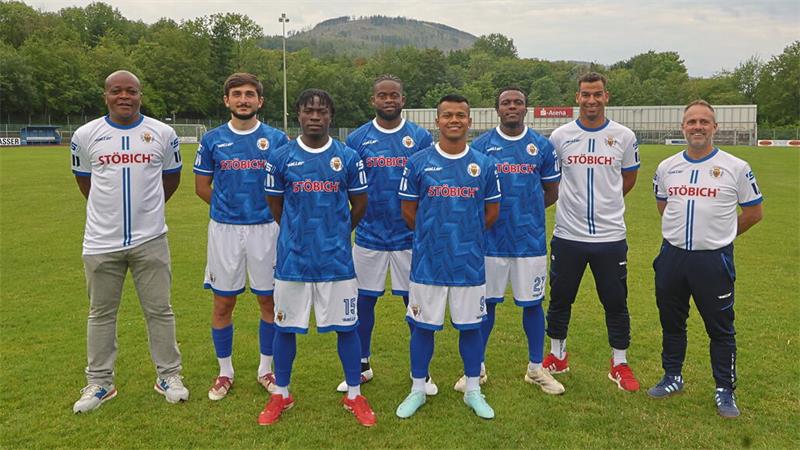 Sieben Männer in Fußballtrikots und Trainingsanzügen stehen nebeneinander auf einem Fußballfeld vor einer Tribüne und bewaldeten Hügeln im Hintergrund.