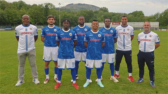 Sieben Männer in Fußballtrikots und Trainingsanzügen stehen nebeneinander auf einem Fußballfeld vor einer Tribüne und bewaldeten Hügeln im Hintergrund.