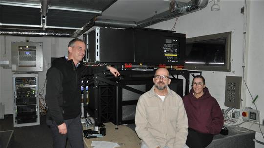 Drei Personen in einem technischen Raum mit großem schwarzem Gerät auf einem Gestell und mehreren elektronischen Geräten an den Wänden.