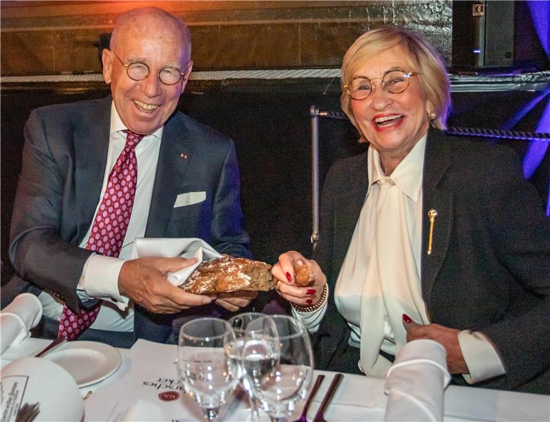 Zwei Personen in formeller Kleidung sitzen an einem gedeckten Tisch und halten gemeinsam ein Stück Brot