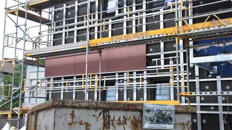 Fassade eines Gebäudes mit Gerüst und brauner Verkleidung, davor ein rostiger Container mit einem Bild darauf.