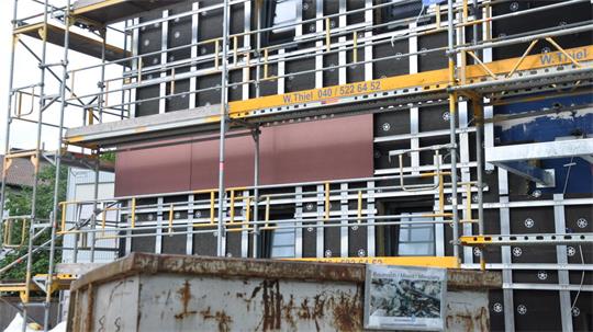 Fassade eines Gebäudes mit Gerüst und brauner Verkleidung, davor ein rostiger Container mit einem Bild darauf.