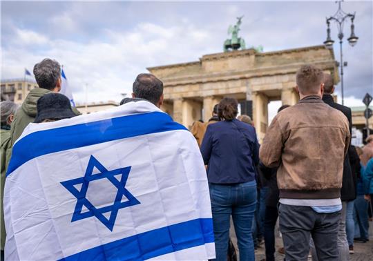 Teilnehmer der Kundgebung „Aufstehen gegen Terror, Hass und Antisemitismus – in Solidarität und Mitgefühl mit Israel“ stehen umhüllt in eine israelische Flagge vor dem Brandenburger Tor in Berlin.