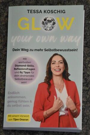Buchcover mit dem Titel 'GLOW your own way' von Tessa Koschig, darunter der Untertitel 'Dein Weg zu mehr Selbstbewusstsein' und ein Foto einer Frau in rotem Blazer vor hellem Hintergrund.
