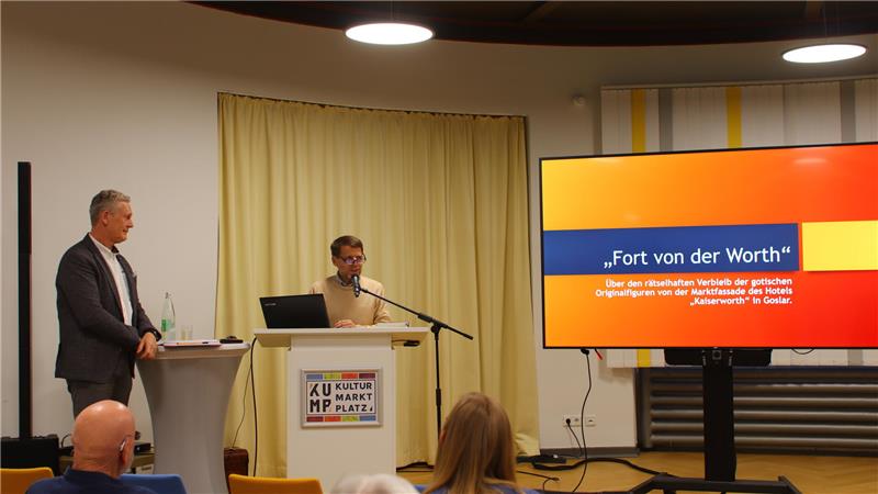 Zwei Personen stehen an einem Rednerpult und einem Stehtisch vor einem Publikum, rechts ein großer Bildschirm mit der Überschrift 'Fort von der Worth'.