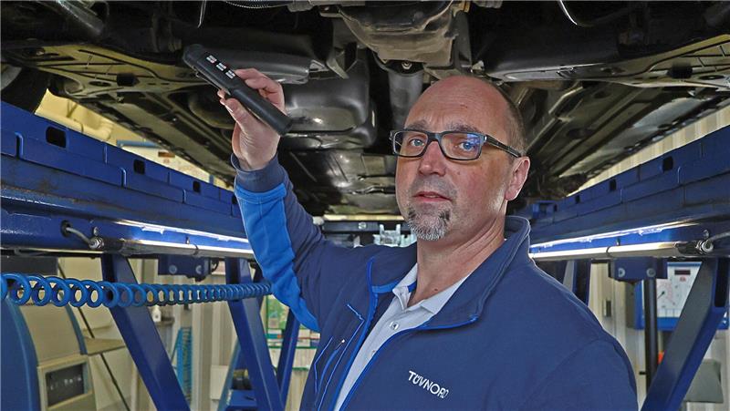 Person in blauer Jacke mit Aufschrift "TÜV NORD" hält ein Messgerät unter ein Fahrzeug auf einer Hebebühne in einer Werkstatt.