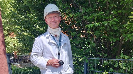 Mann in weißer Arbeitskleidung und Helm steht vor grünem Gebüsch und hält eine Lampe in der Hand.