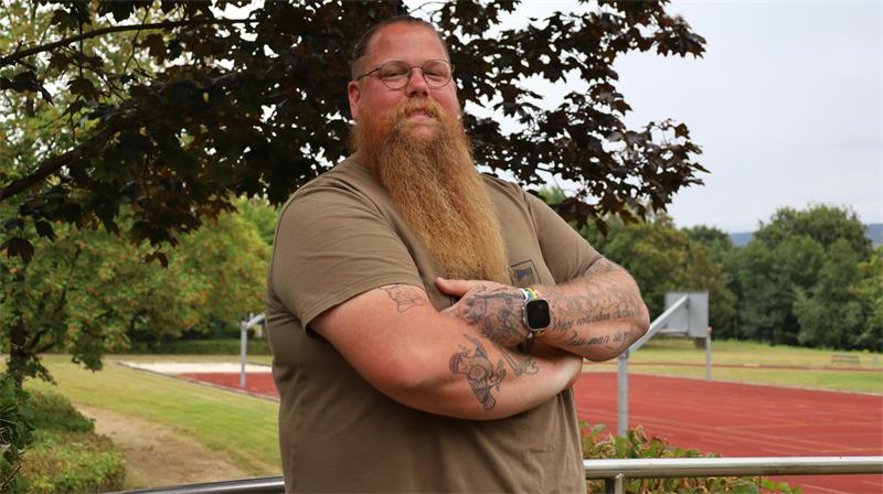 Mann mit langem Bart, Brille und Tattoos steht mit verschränkten Armen vor einem Sportplatz und Bäumen