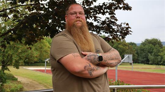 Mann mit langem Bart, Brille und Tattoos steht mit verschränkten Armen vor einem Sportplatz und Bäumen