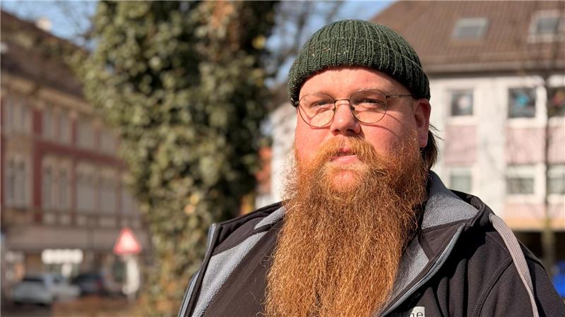 Mann mit langem roten Bart, schwarzer Kapuzenjacke und grünem Armband steht im Freien vor Gebäuden und Bäumen