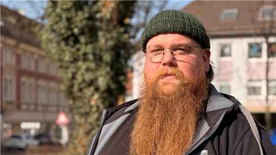 Mann mit langem roten Bart, schwarzer Kapuzenjacke und grünem Armband steht im Freien vor Gebäuden und Bäumen