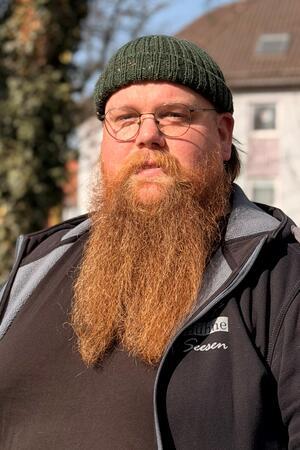 Person mit langem roten Bart, grüner Strickmütze und schwarzer Jacke mit Aufschrift.