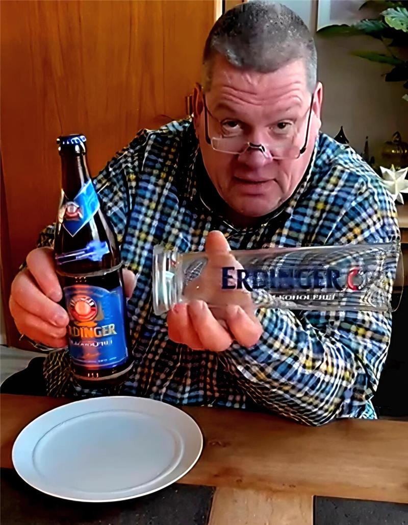 Person in kariertem Hemd hält eine Flasche Erdinger Weißbier und ein Erdinger-Glas über einem gedeckten Holztisch mit weißem Teller.