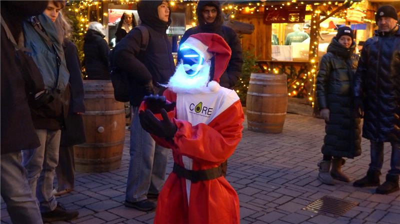 Weihnachtsmann mit leuchtender, blauer LED-Maske und rotem Kostüm steht auf gepflastertem Platz mit weihnachtlicher Beleuchtung und mehreren Personen im Hintergrund.