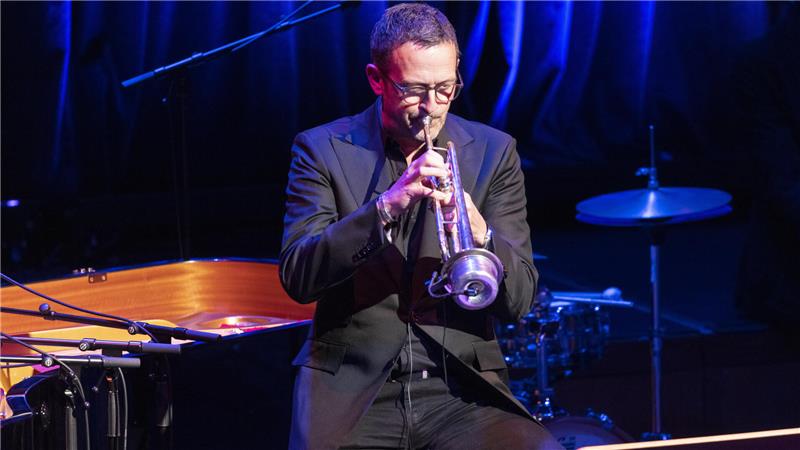 Jazz-Trompeter Till Brönner komplettiert Pfalzkonzerte Line-up Mann in dunklem Anzug spielt Trompete auf Bühne mit Schlagzeug und Klavier im Hintergrund.