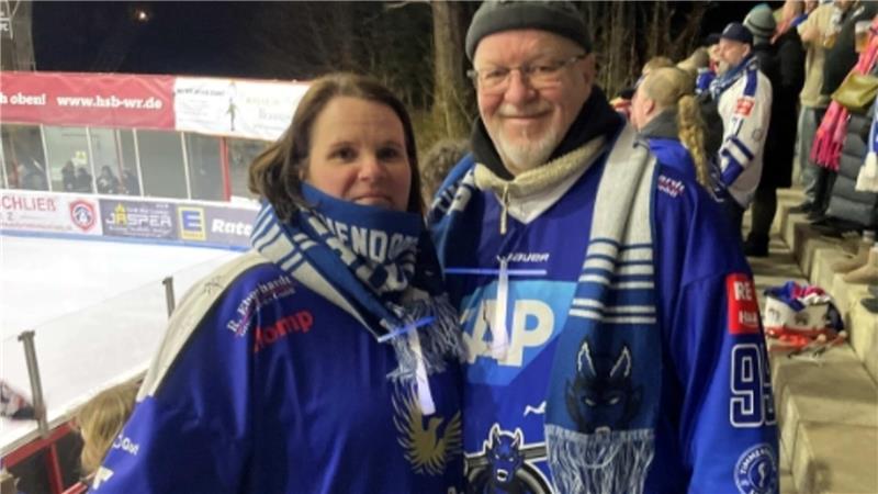 Zwei Personen in blauen Eishockeytrikots und passenden Schals stehen nebeneinander vor einer Tribüne mit Zuschauern.