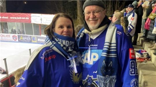 Zwei Personen in blauen Eishockeytrikots und passenden Schals stehen nebeneinander vor einer Tribüne mit Zuschauern.