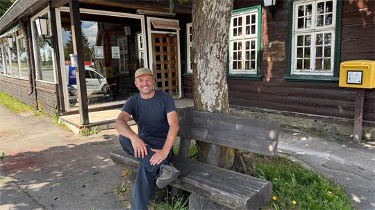 Das Foto zeigt einen Mann, der auf einer Bank vor einem Haus sitzt.