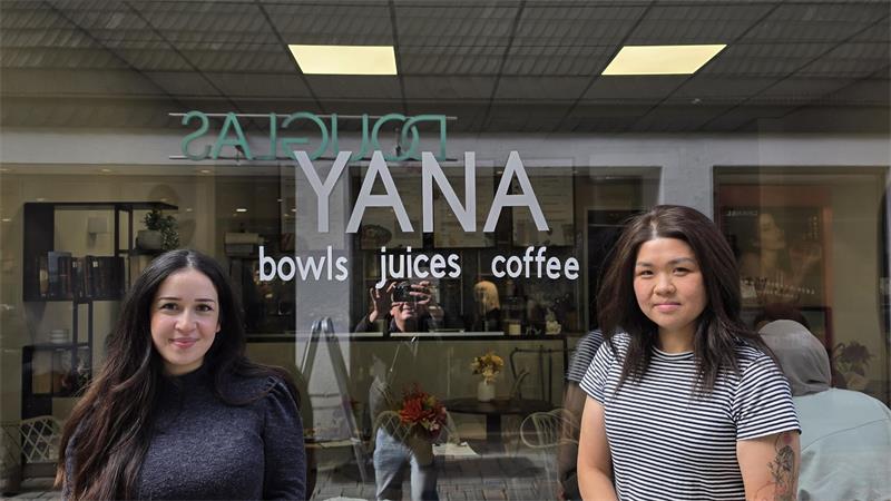 Zwei Frauen stehen vor einem Caféfenster mit der Aufschrift 'YANA bowls juices coffee'