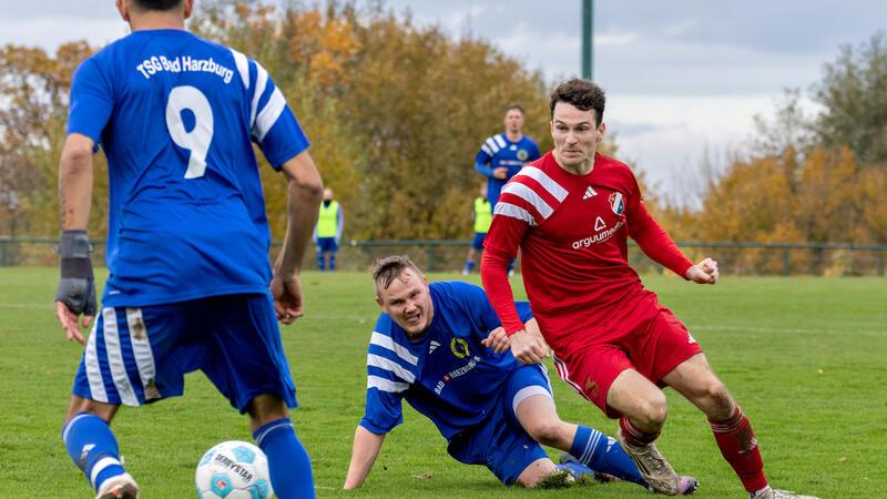 Fußballspieler in blauen und roten Trikots kämpfen auf einem Spielfeld um den Ball, einer der Spieler in Blau liegt am Boden
