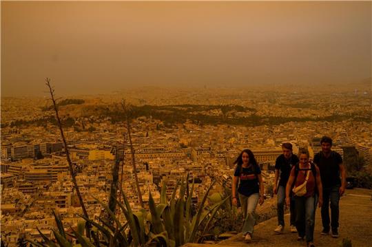 Touristen spazieren auf dem Lycabettus-Hügel, während der Himmel über Athen mit dem antiken Akropolis-Hügel im Hintergrund infolge von Saharastaub eingetrübt ist.