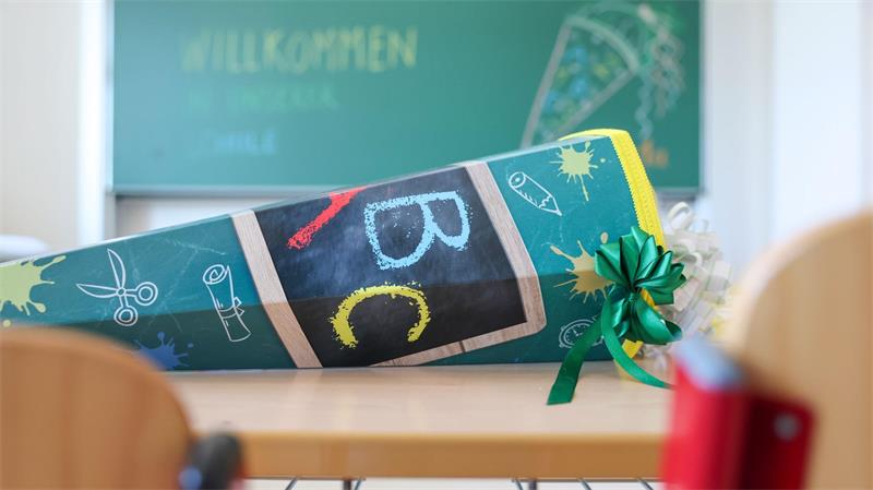 Schultüte mit ABC-Motiven und grünem Schleifenband auf einem Schultisch vor einer Tafel mit der Aufschrift 'WILLKOMMEN'