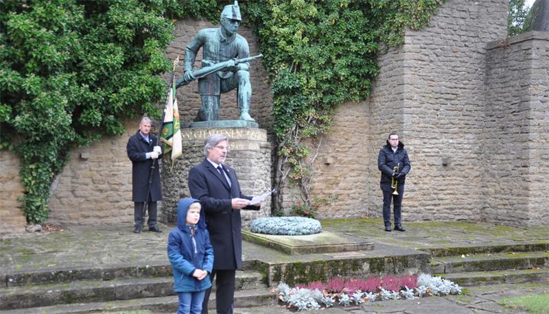 Mann hält Rede vor Kriegerdenkmal mit Soldatenskulptur, Kind steht daneben, zwei weitere Personen halten Fahne und Trompete in einer Gedenkstätte mit Steinmauer und Pflanzen.