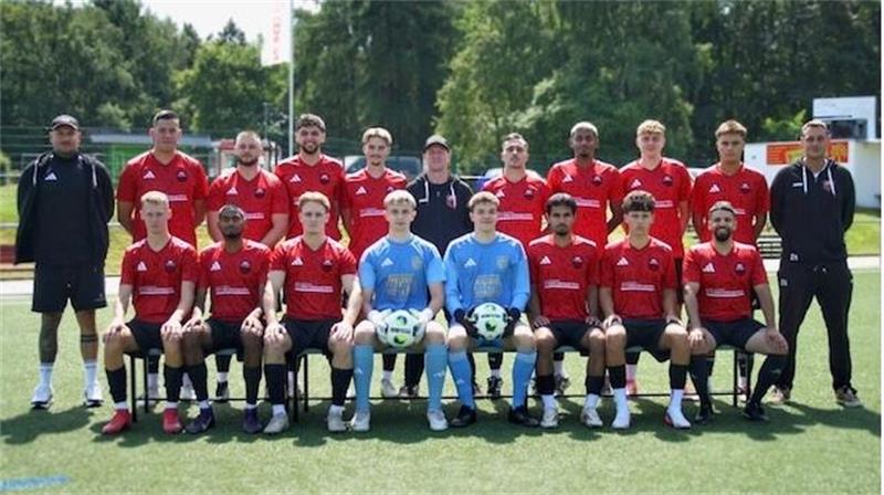Mannschaftsbild einer Fußballmannschaft mit Spielern in roten Trikots und zwei Torhütern in blauen Trikots auf einem Fußballfeld vor Bäumen