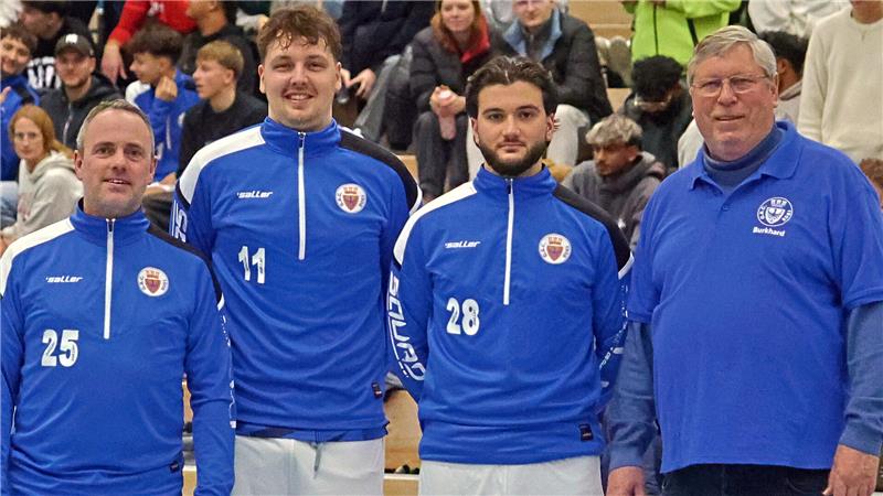 Vier Männer in Sportkleidung stehen nebeneinander in einer Halle, drei tragen blaue Trainingsanzüge mit weißen Hosen, einer trägt ein blaues Poloshirt und Jeans.