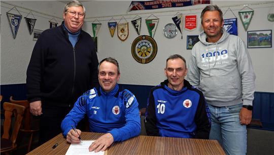 Vier Männer in einem Raum mit Wimpeln an der Wand zwei sitzen am Tisch einer unterschreibt ein Dokument zwei stehen dahinter.