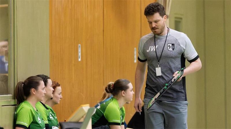 Trainer mit Sportkleidung und Pfeife steht vor sitzenden Spielerinnen in grünen Trikots in einer Halle