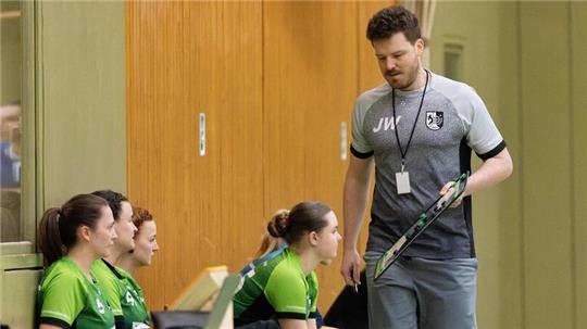 Trainer mit Sportkleidung und Pfeife steht vor sitzenden Spielerinnen in grünen Trikots in einer Halle