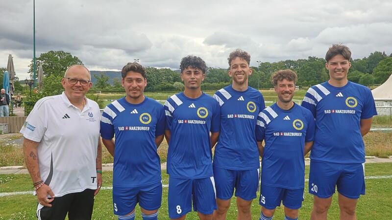 Fünf Männer stehen nebeneinander auf einer Wiese, vier tragen blaue Fußballtrikots mit gelbem Logo und Nummern, der fünfte trägt ein weißes Poloshirt mit Aufdruck.