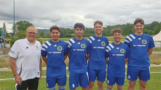 Fünf Männer stehen nebeneinander auf einer Wiese, vier tragen blaue Fußballtrikots mit gelbem Logo und Nummern, der fünfte trägt ein weißes Poloshirt mit Aufdruck.