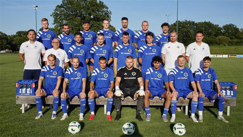 Mannschaftsbild einer Fußballmannschaft mit Spielern in blauen Trikots und vier Personen in weißen Poloshirts auf einer Wiese vor Bäumen
