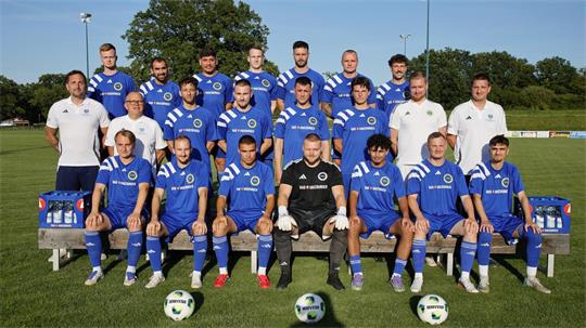 Mannschaftsbild einer Fußballmannschaft mit Spielern in blauen Trikots und vier Personen in weißen Poloshirts auf einer Wiese vor Bäumen