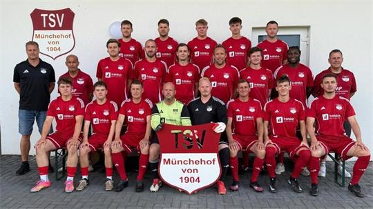 Mannschaftsbild einer Fußballmannschaft mit 20 Spielern und zwei Trainern vor einer weißen Wand, die Spieler tragen rote Trikots und halten ein Schild mit der Aufschrift 'TSV Münchehof von 1904'