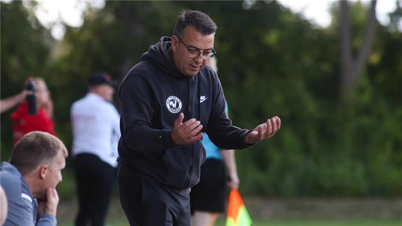 Mann in schwarzer Nike-Sportkleidung gestikuliert auf einem Fußballfeld mit unscharfem Hintergrund und weiteren Personen