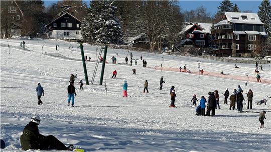 Das Foto zeigt einen sitzenden Snowboardfahrer im Vordergrund, einen vollen Skihang mit vielen Skifahrern und Passanten.