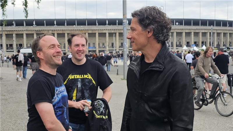 Drei Männer in T-Shirts und Jacke stehen im Freien vor einem großen Gebäude mit Säulen und sprechen miteinander