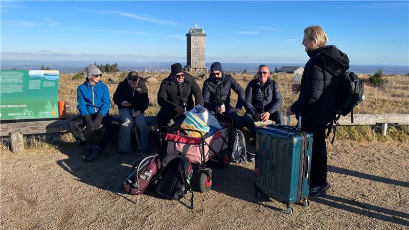 Fünf Personen sitzen auf einem Baumstamm und eine Person steht vor ihnen. In der Mitte steht ihr Gepäck.